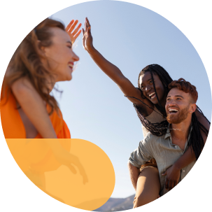 A group of friends laughing and high-fiving on a sunny day, with one woman on a man’s back and another reaching out.