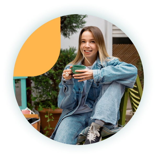 A young woman in a denim outfit smiles while holding a green coffee cup, sitting casually in a cozy outdoor setting