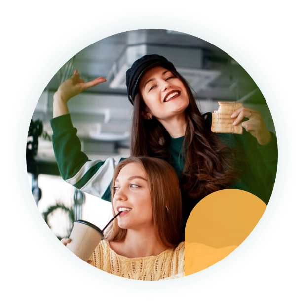 two young women in casual clothes smile and hold drinks in a café, one raising her arm in a joyful pose.