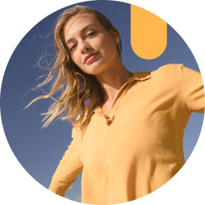 A woman in a mustard yellow long-sleeve shirt poses confidently under a clear blue sky with wind in her hair.