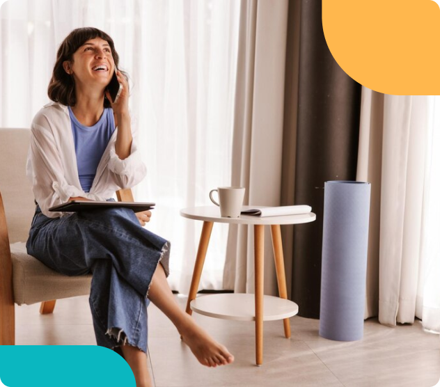 A woman sits barefoot in a bright living room, laughing on the phone with a tablet in her lap and a coffee mug nearby.