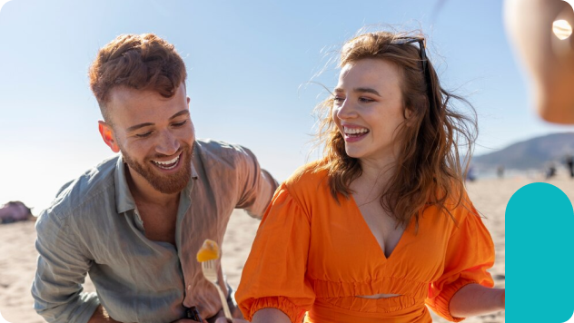 A man and woman share a laugh at the beach, the man holding a fork with fruit and the woman wearing a bright orange dress.