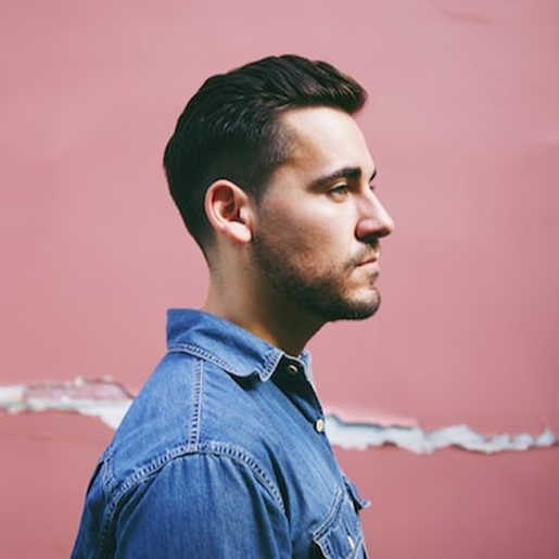 A man in a denim shirt stands in profile against a pink wall with a torn white stripe across it.