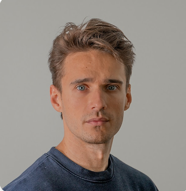 A man with tousled hair wears a navy crewneck sweatshirt and looks directly at the camera against a plain grey background.