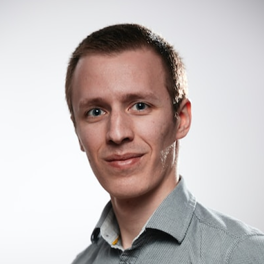 A man in a light grey button-up shirt smiles slightly at the camera against a neutral studio backdrop.
