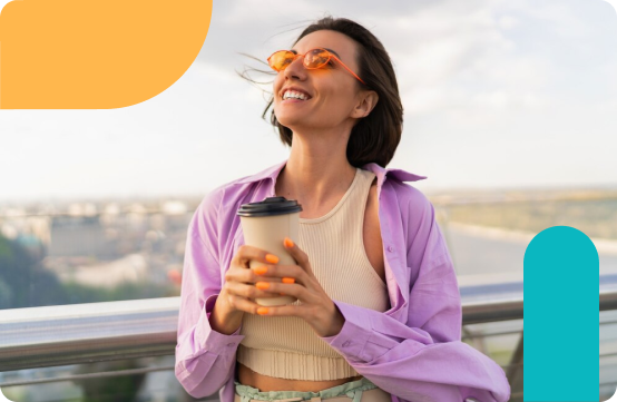 A smiling woman in orange sunglasses and a lavender shirt holds a coffee cup while standing on a rooftop with a city view.