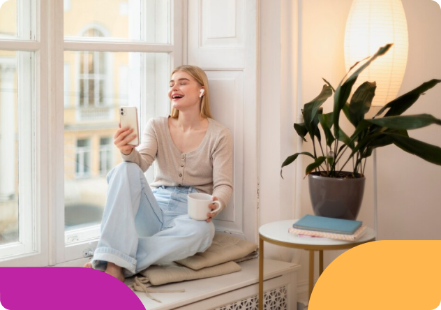 A woman sits on a window seat with a coffee mug, smiling during a video call on her phone while wearing wireless earbuds.