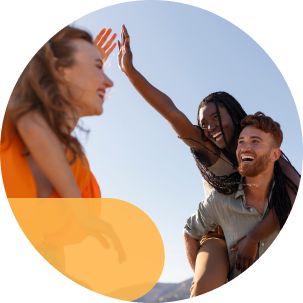 A group of friends laughing and high-fiving on a sunny day, with one woman on a man’s back and another reaching out.