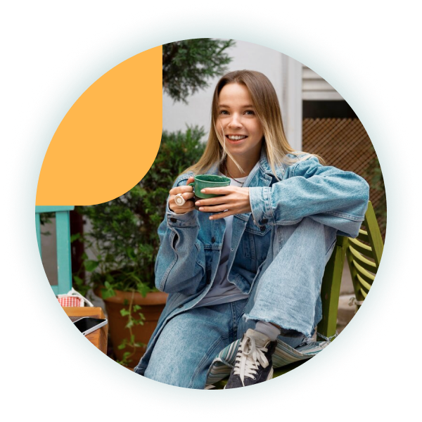 A young woman in a denim outfit smiles while holding a green coffee cup, sitting casually in a cozy outdoor setting