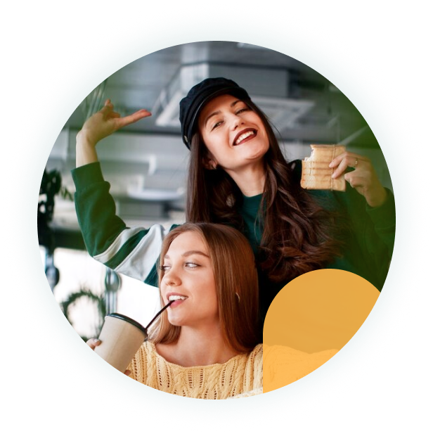 two young women in casual clothes smile and hold drinks in a café, one raising her arm in a joyful pose.