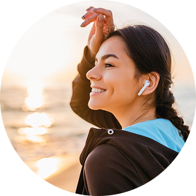 A woman smiles while wearing wireless earbuds, standing on the beach during sunset with her hand brushing back her hair.