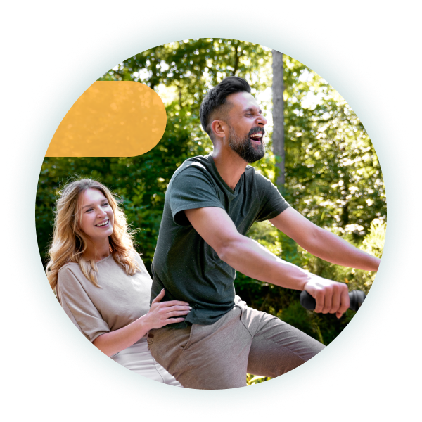 A couple laughs as they ride a bike together through a forested path, with the woman sitting behind the man who’s steering.