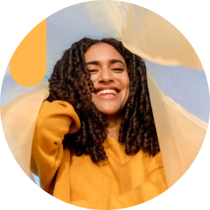 A cheerful woman in a yellow top smiles as soft, sheer fabric flows around her against a blue sky background.
