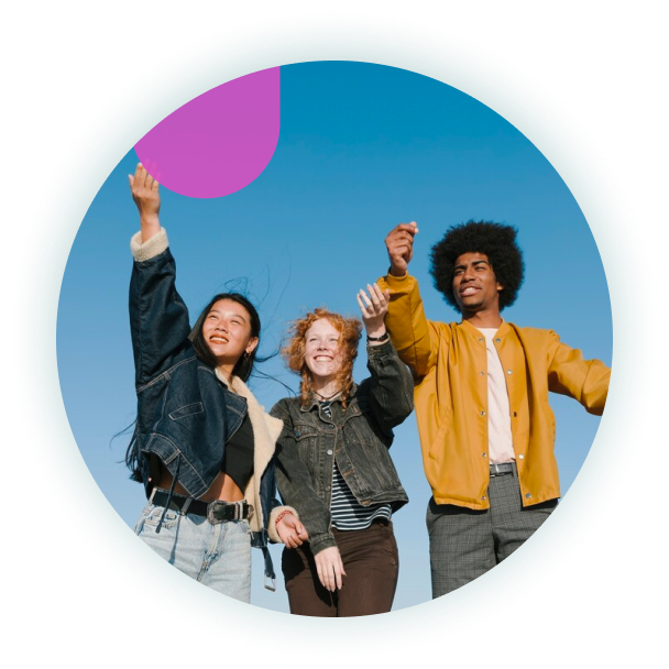 Three friends stand outdoors under a clear blue sky, smiling and reaching upward with joy and energy.