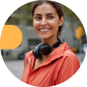 A woman in an orange jacket smiles while wearing black headphones around her neck, standing on a city street